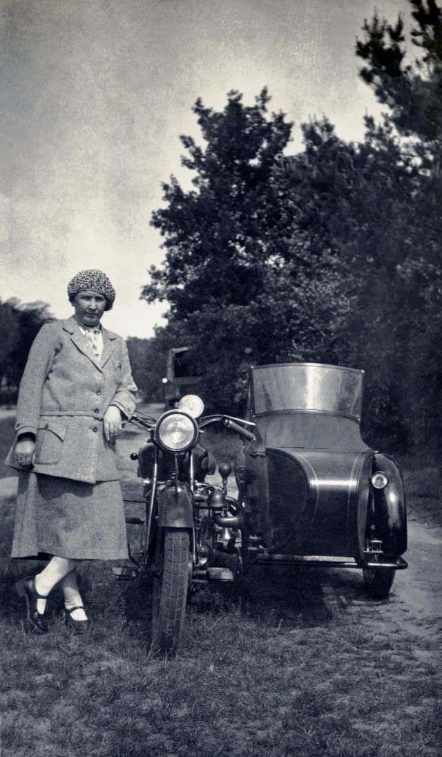 A stylish lady in a female suit posing with a sidecar motorcycle in the countryside, circa 1930s.