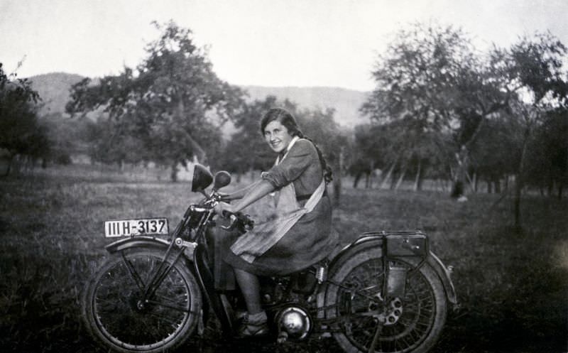 A young lady in a dress and apron posing in the saddle of an ancient motorcycle. The print is dated "september 1931" on reverse, the bike is registered in the german state of württemberg.