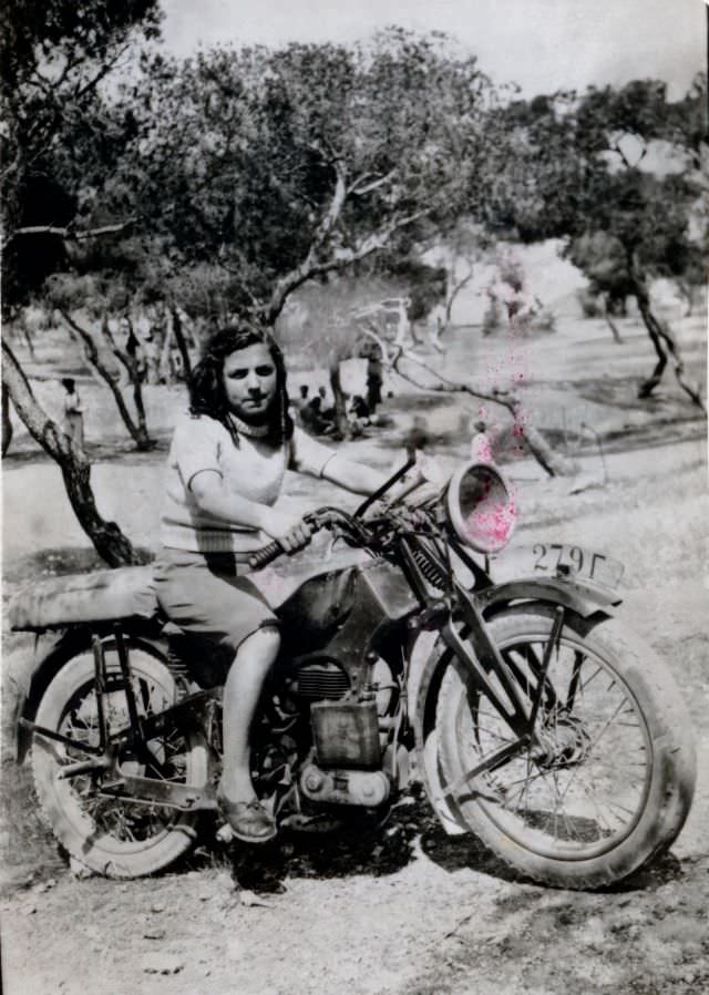 A teenage girl posing in the saddle of an ancient motorcycle in the countryside. The location seems to be an olive grove, somewhere in rural greece, circa 1950s.