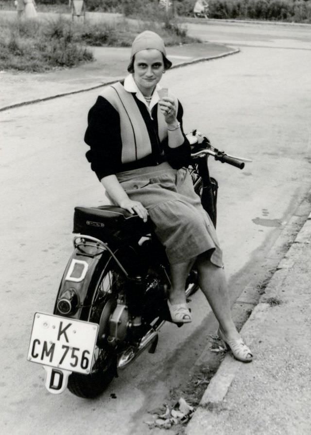A young lady with an ice cream cone in her hand posing in the saddle of a bmw r25/2. The bike is registered in the west german city of cologne, circa 1956.