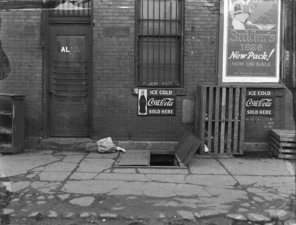 21st and Chestnut, 1930 - View of an open cellar door behind a grocery store. Advertising for Coca-Cola and Snider's ketchup are hung on the building.