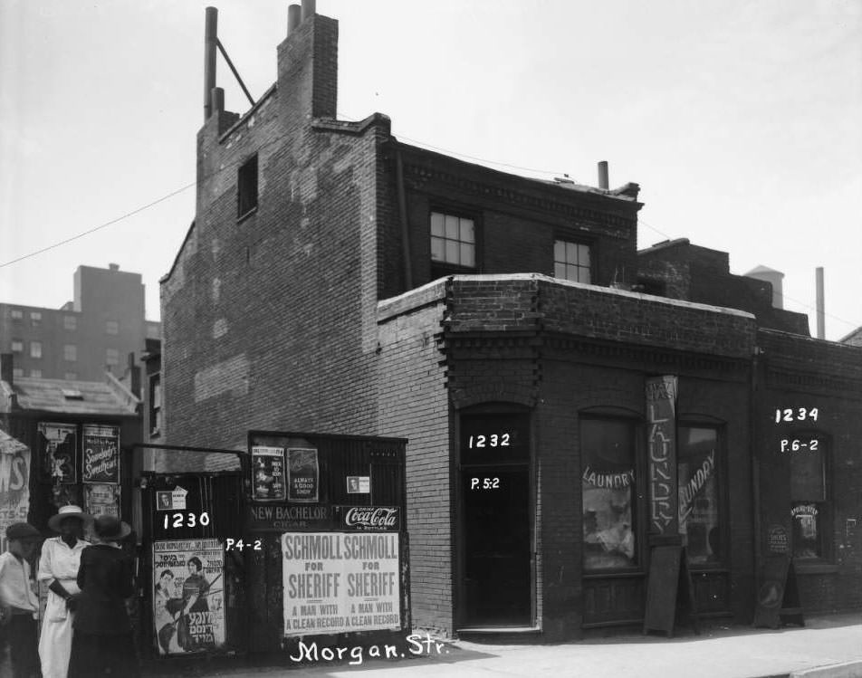 Exterior view of a laundromat and shoe repair shop with three people standing in front of billboards. 1232 and 1234 Morgan St, 1930