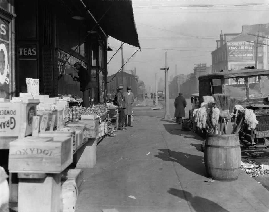 View of Molasky Grocery Store near the intersection of N. 13th and O'Fallon St. with a barrel of brooms and a man painting on the window, 1930