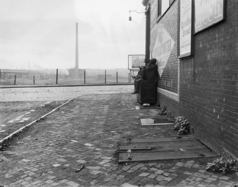 View of two men sitting on a cupboard beside a building at Leonard Street and Market Street. Blue Valley Butter sign is visible, 1930