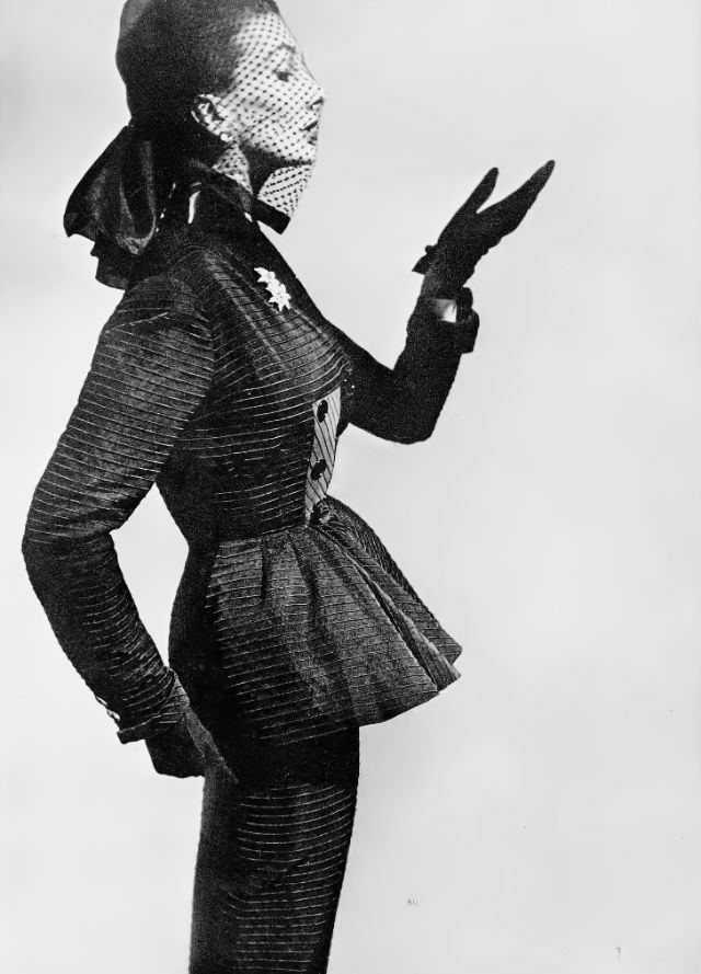 Barbara Goalen in elegant suit of black ottoman silk with a raised horizontal rib and a front-flaring basque on the long jacket by Lachasse, worn with silk spotted veil, 1951.
