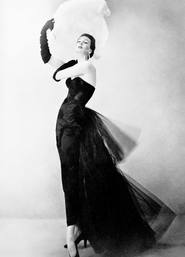 Barbara Goalen in sleek black satin sheath with floating train of tulle in back, the satin is embossed with velvet and ostrich by Mattli, 1951.
