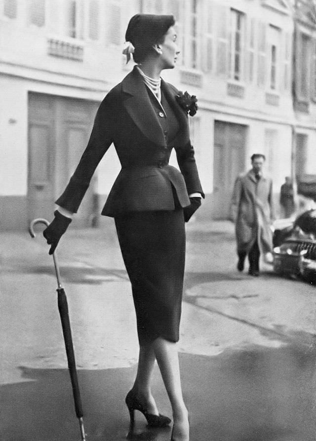 Barbara Goalen in fitted navy blue wool suit with horseshoe neckline filled in with matching vest, skirt is made up of two petal-shaped pieces, by Christian Dior, 1950.