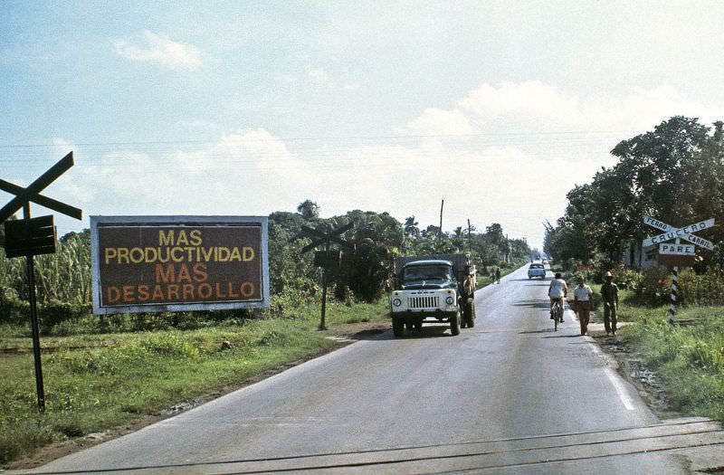 Country road in the interior of the island. Warning over rail crossing, truck and people passing. A billboard recommends "More productivity and More development", Cuba, 1976