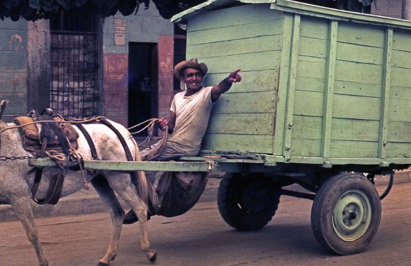 Man sitting in his green wagon pulled by a horse, laughing and pointing a direction with his arm, Cuba, 1976