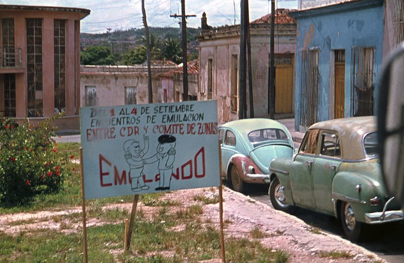Old American cars parked on a street with old houses. In a sign at the street: "Emulation Meetings between CDR and Zoning Committee announced", Cuba, 1976