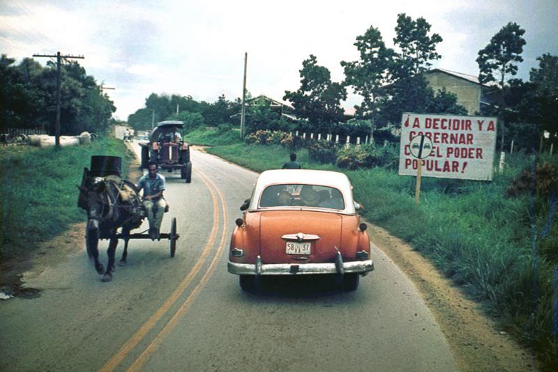 On the road, an old American car, a horse and cart and a tractor. Sign near the road of socialist government message: "To decide and govern with the people's power", Cuba, 1976