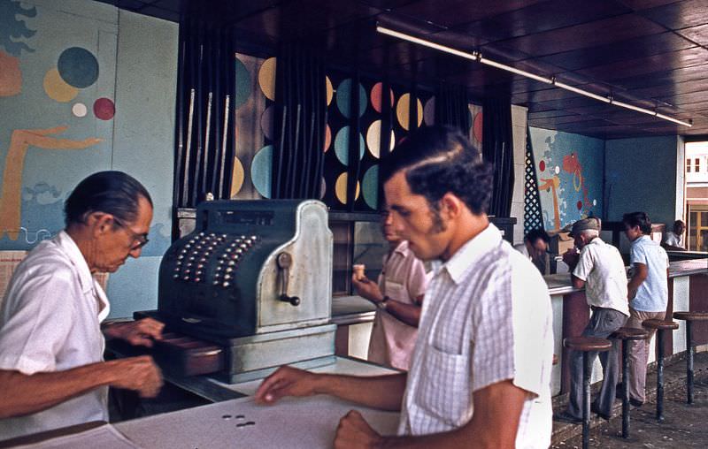 Cuba, Havana, 1976. In a bar, a customer pays their consumption to the cashier who works with an old cash machine