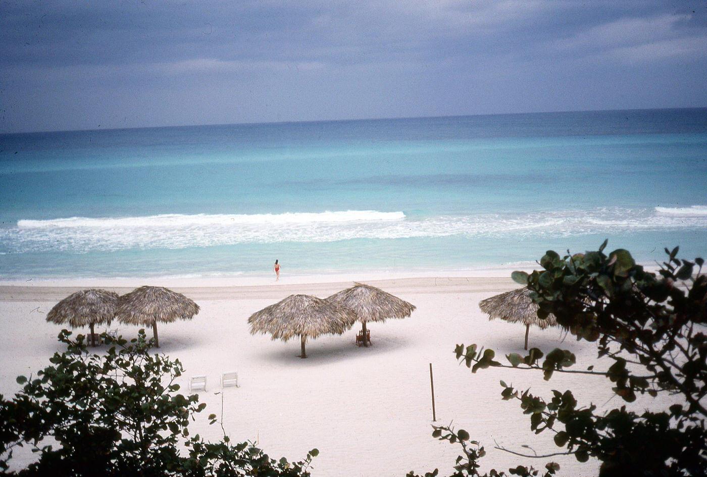 Deserted beach at an unidentified resort where one woman in a red swimsuit walks into the surf, Varadero, Cuba, January 1983.