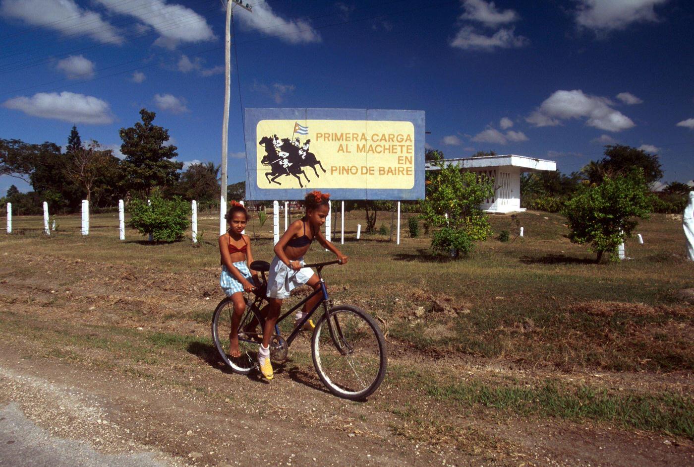 Two Cuban girls riding bicycles with a poster commemorating a historical event of the Cuban War of Independence, Cuba, 1990.
