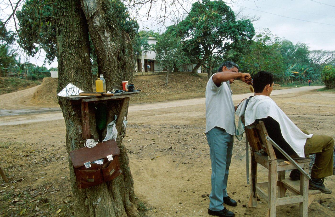 Cuban man combing a Cuban boy's hair beside a tree trunk, Cuba, 1990.