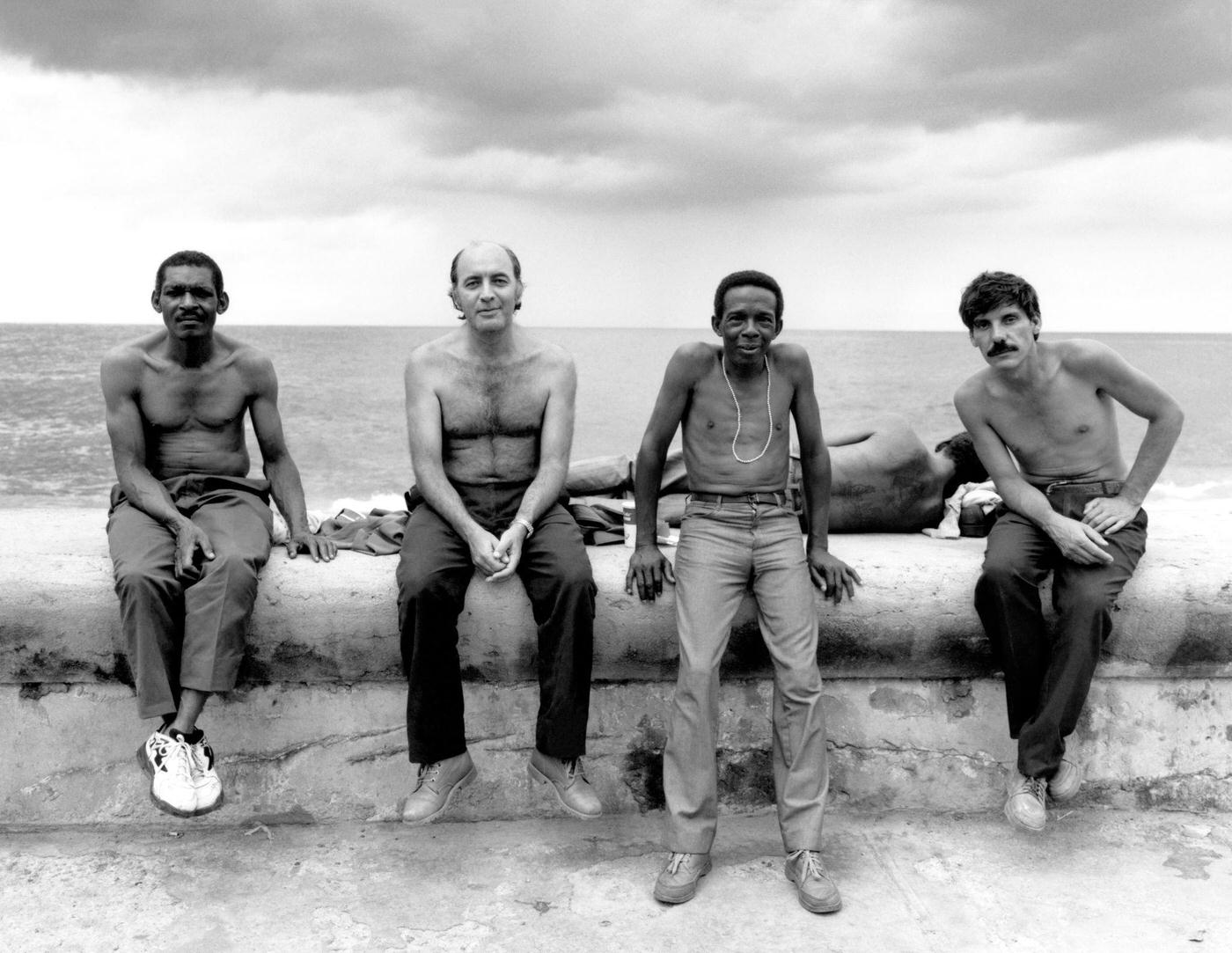 General view of four shirtless men posing for a portrait while sitting on a wall in Havana, Cuba, 1996.