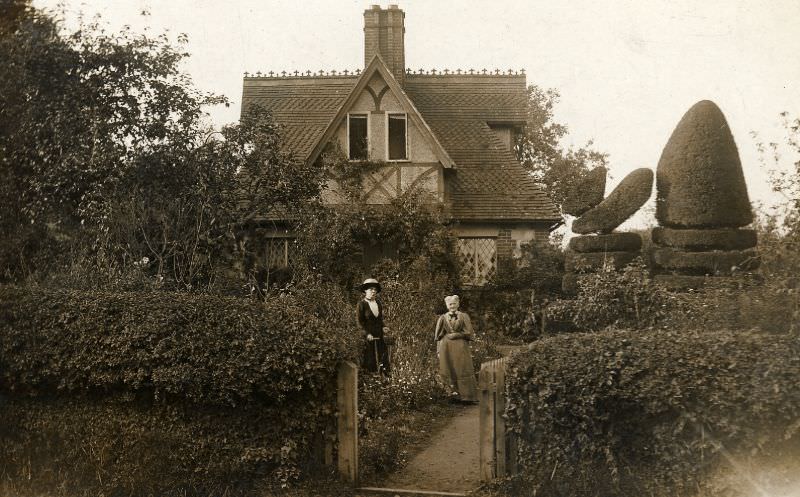 Two women stand in the garden of a Victorian or Edwardian house built in a romantic cottage style. The garden is well tended and includes examples of topiary