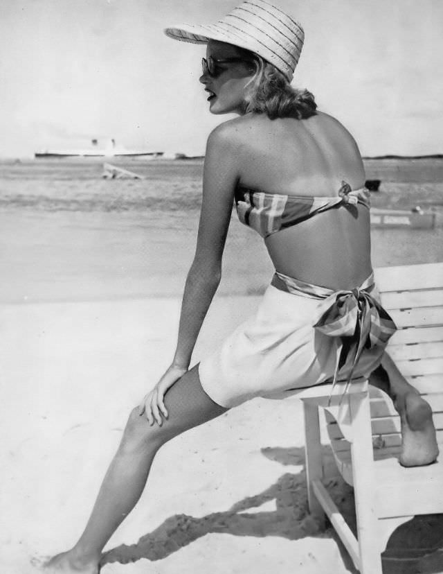 Model in cotton halter top, plaided in yellow, black and brown with plain shorts of yellow sharkskin, tied with plaid sash, by Cabana Sportswear, Montagu Beach, Nassau, Bahamas, Harper's Bazaar, 1948.