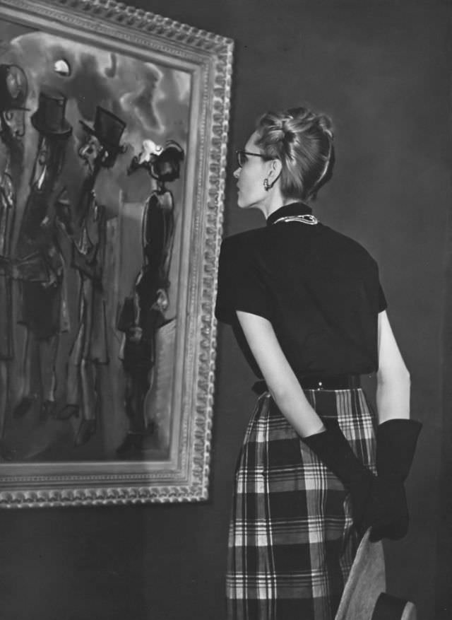 Model in a bright-and-black gingham dress with bold plaid skirt and short sleeves, belted in black patent leather by David Crystal, in front of a painting by Max Weber, Vogue, 1945.