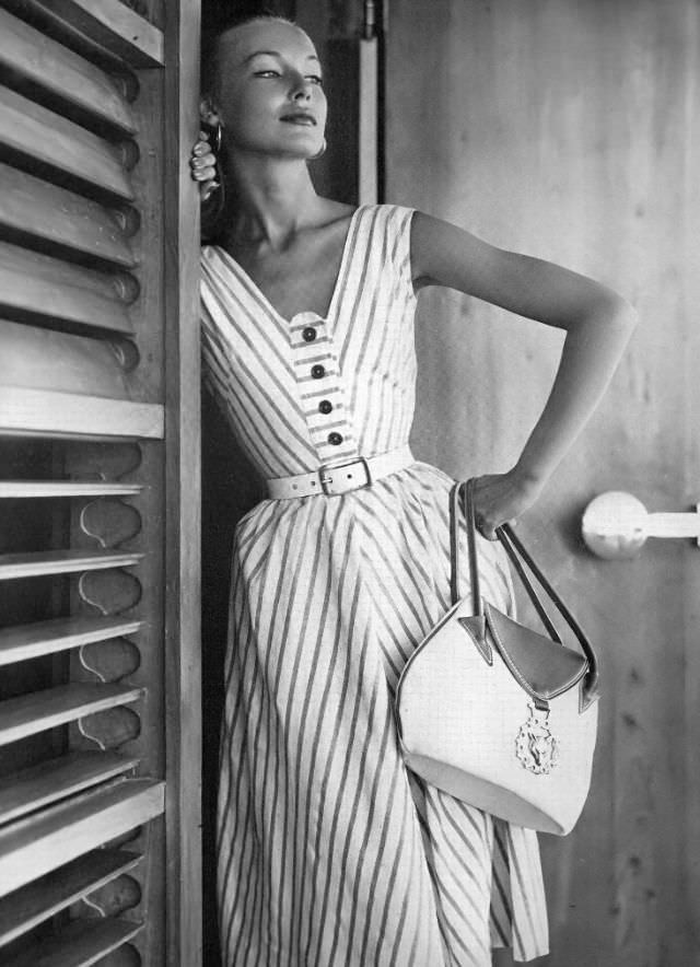 Liz Pringle in pretty sundress of brick-red stripes on white denim, low in front and in back by Sailing Blues, white belt by Calderon, handbag by Van S, Jamaica, Vogue, May 1, 1953