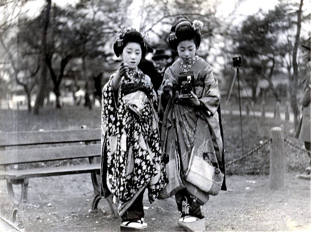 A Maiko girl taking a photograph with an early Kodak folding camera, while being guided by another Maiko, 1920s