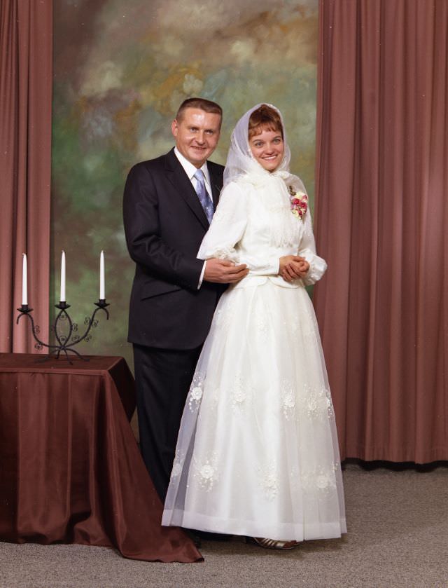 Wedding portrait of a Doukhobor couple. The groom is wearing a dark suit, with a white shirt and blue tie. The bride is wearing a white dress, a corsage, and a traditional white headscarf, November 27, 1971