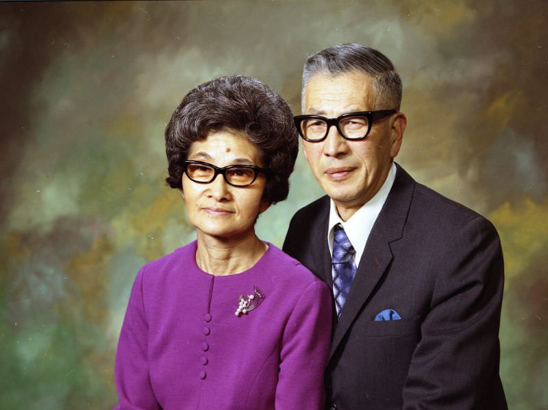 Mr and Mrs Kinoshita. Mrs Kinoshita is wearing a purple top with a metal pendant. Mr. Kinoshita is wearing a white shirt, blue-silver tie, and a charcoal suit jacket. Both individuals are wearing black-framed glasses, February 1973