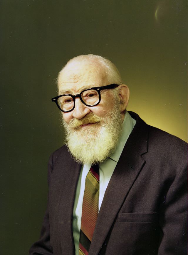 Portrait of an old man wearing a dark grey suit jacket over a white shirt and diagonally-striped tie. He has a large white beard and black-framed glasses, September 10, 1975