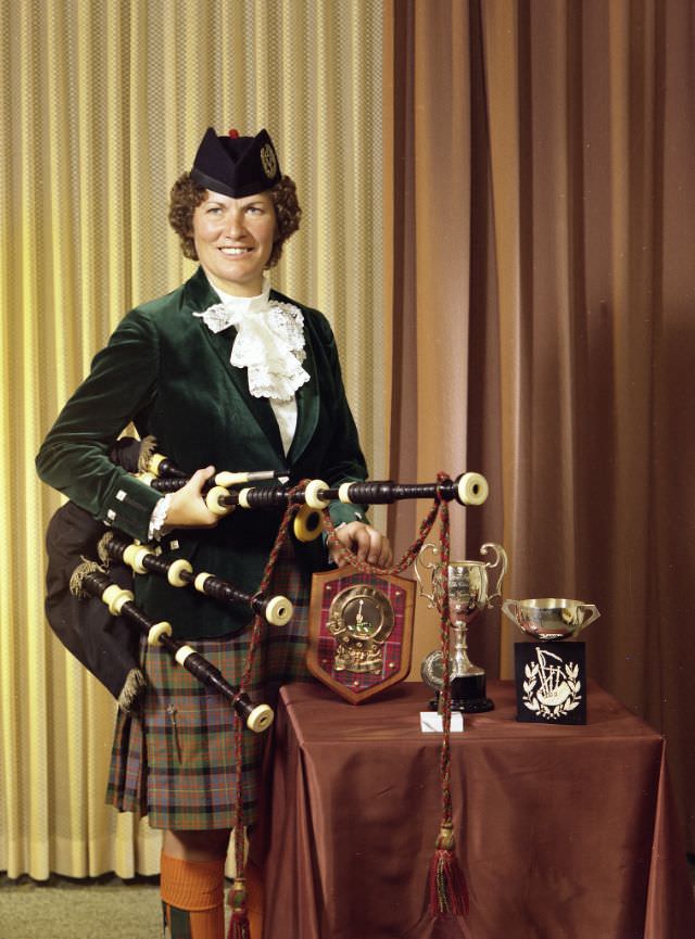 Portrait of a woman dressed in traditional Scottish attire, posing with three trophies. The woman is wearing a black hat and is holding a set of bagpipes in her right hand, June 22, 1977