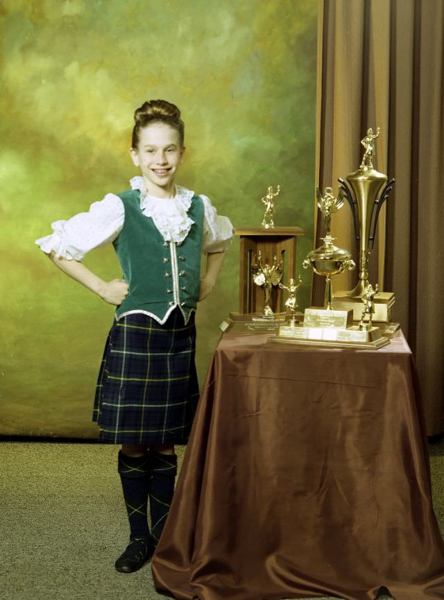 Portrait of a girl posing with three trophies. The girl is dressed in a Highland dance outfit and the trophies have been placed on a low, draped table on the right-hand side of the image, March 29, 1978