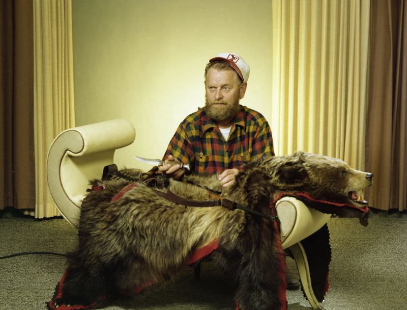 Portrait of a man with a bear hide and hunting paraphernalia. He is wearing a cap and plaid button-up shirt, June 3, 1978