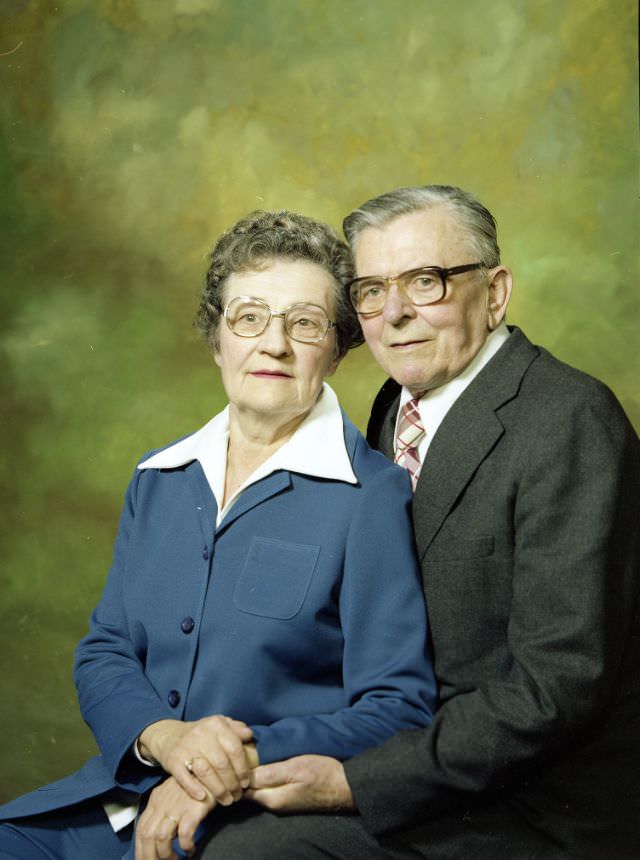 Portrait of an older couple. The woman is wearing a blue pant suit over a white shirt with a broad collar. The man is wearing a charcoal grey suit over a white shirt and plaid tie. Both individuals are wearing glasses, November 22, 1978