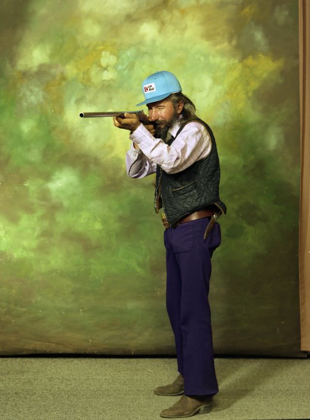Portrait of a man in purple pants, a turquoise cap, and a long-sleeved light shirt. He is holding a gun, and standing before a green background, 1979