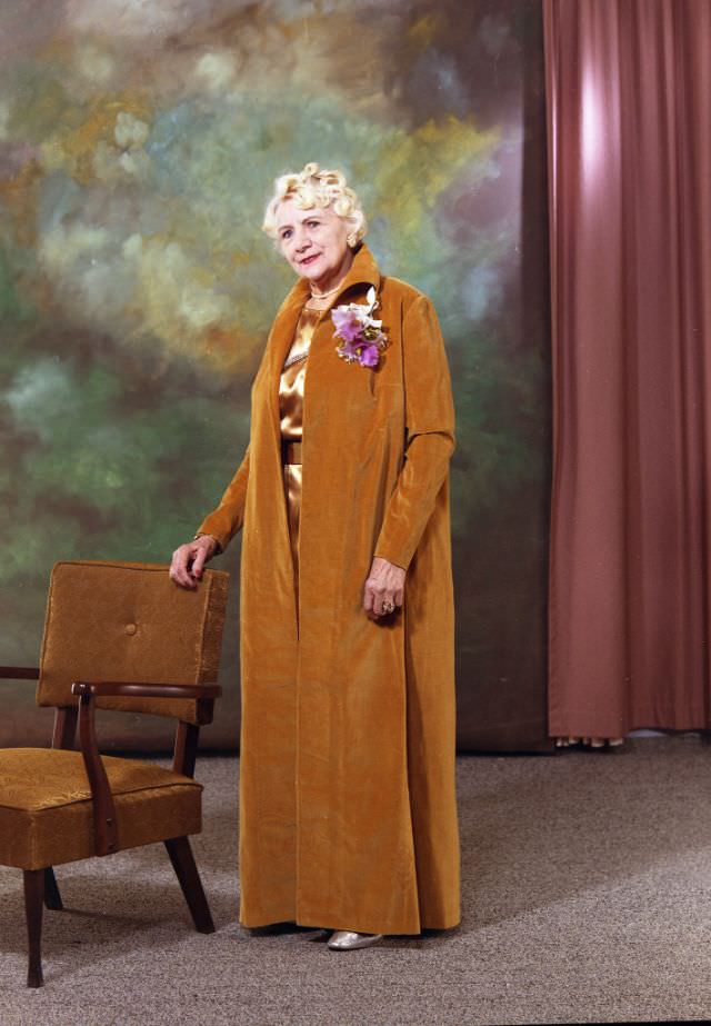Portrait of Kitty Gilbert who dressed in a floor-length gold velvet evening jacket that has been adorned with a corsage of purple flowers, January 28, 1971