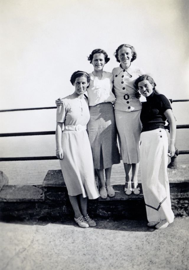 A company of four well-to-do ladies posing on a promenade overlooking the sea.
