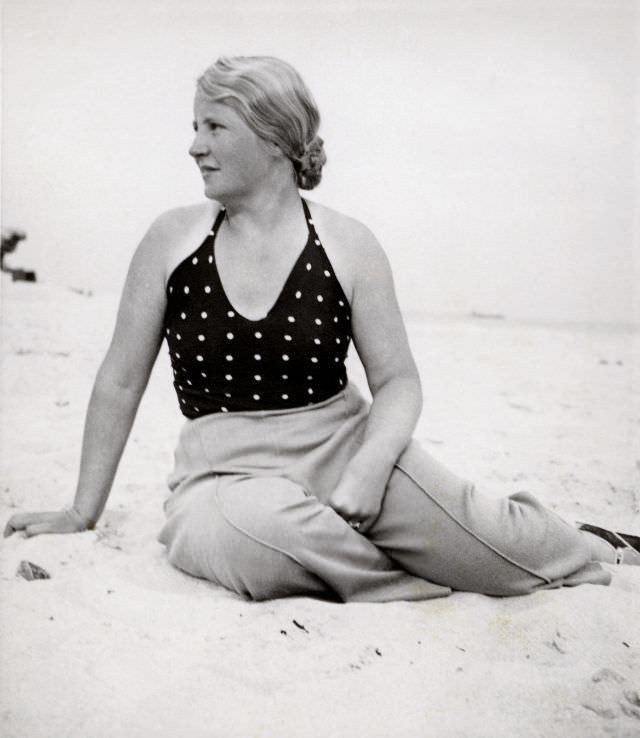 A blonde lady posing on a beach in summertime. She is wearing a polka dot top and slacks with sewn-in creases, 1938