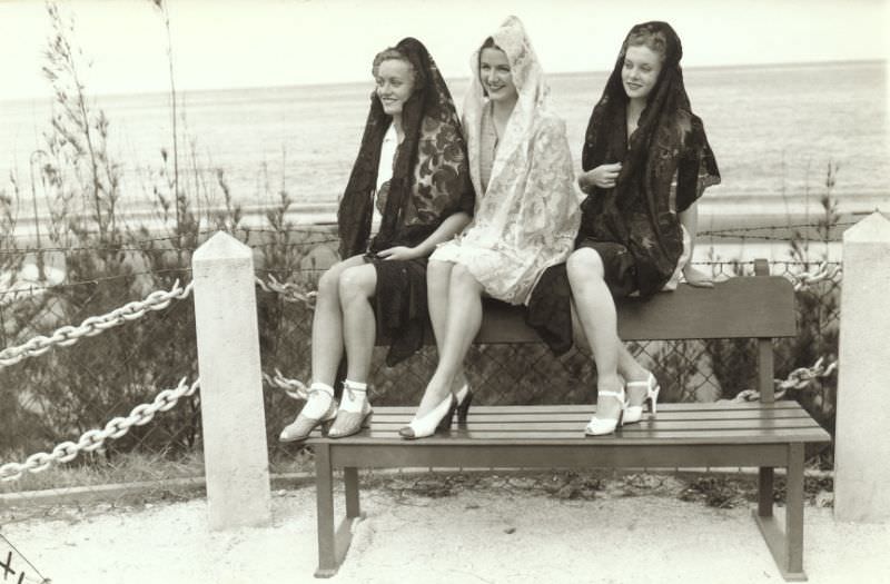 Three young American women posing on a bench at the seaside. They are dressed in traditional Spanish lace Mantillas, 1941