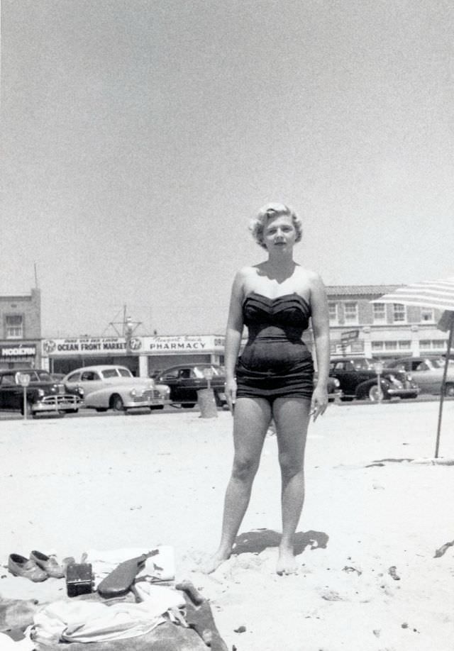 A platinum-blonde lady in a one-piece swimsuit posing on a sunny beach.