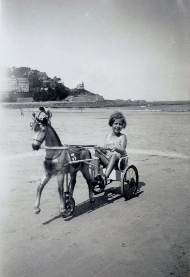 A blonde girl posing in the seat of a pedal tricycle on a beach in midday sunshine.