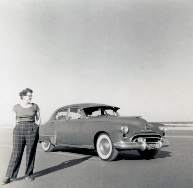 A curly-haired lady dressed in tartan check trousers posing with a 1949 Oldsmobile 88 4-Door Sedan in late afternoon sunshine، 1952