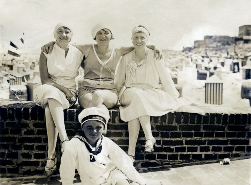 Three fashionable ladies and a freckled boy in a sailor suit posing at the seaside. In the background, the breach is covered with typical German beach chairs (Strandkörbe), 1928