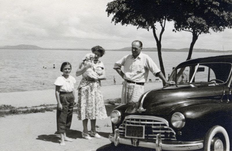 Three members of a middle-class family posing with a Morris Oxford in bright midday sunshine at the seaside, 1955