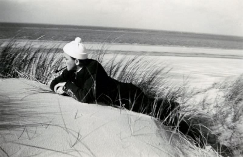 A dapper, pipe-smoking fellow posing in the dunes at the seaside, 1950s