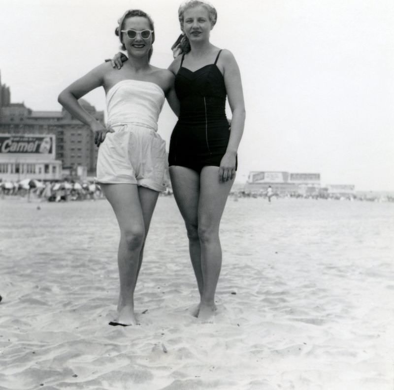 Two cheerful ladies posing on a sandy beach on an overcast day, 1950s