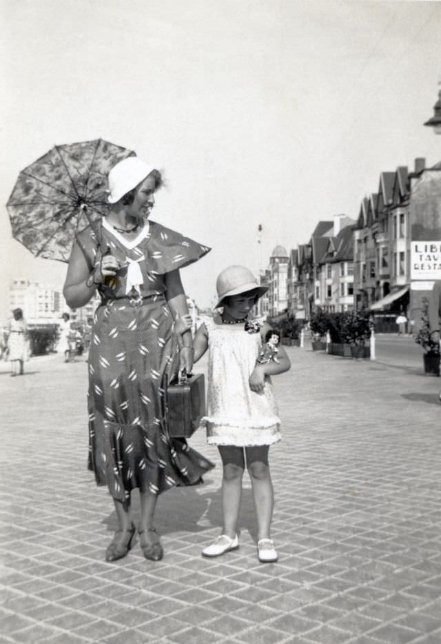 A fashionable lady carrying a lace parasol and her daughter, holding a doll in her arm, posing on a seaside boulevard in summertime, 1930s