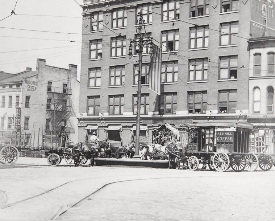 Numerous teams of horse-drawn carriages resting at a watering trough in front of the Kroger Ames James Grocer Co. building at Third Street and Washington in 1920