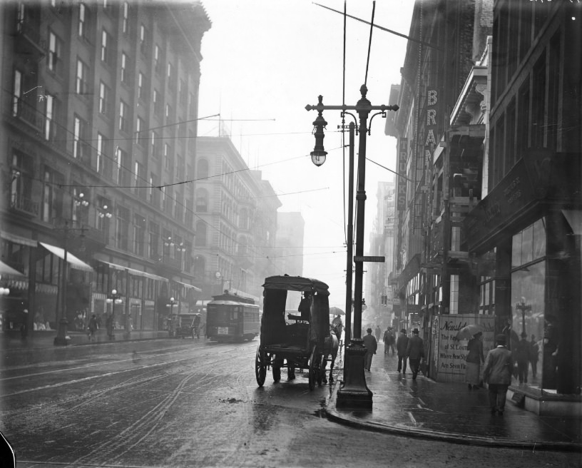 Corner of Seventh Street and Washington Avenue in the rain, with pedestrians walking down the sidewalk and vehicles in the street, 1920s
