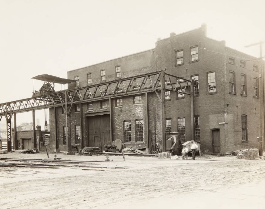 Small factory or warehouse on First Street with an apparatus for loading materials onto rail cars attached to the front of the building and two workmen relaxing in front of it, 1920