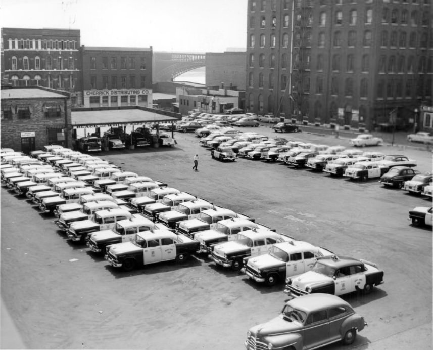 Fleet of taxis parked outside the Black and White Cabs company. Eads Bridge in the background, 1956