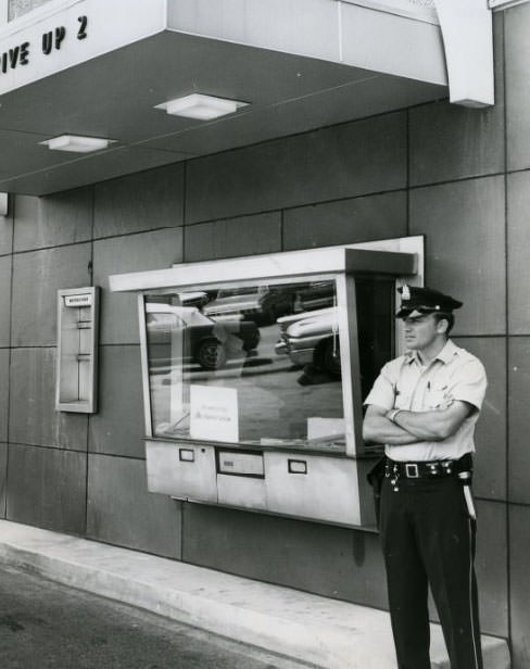 Patrolman stands guard at walk up window of the Gateway Bank, 3412 Union where two holdup men held Peggy Squires, bank teller, 1960